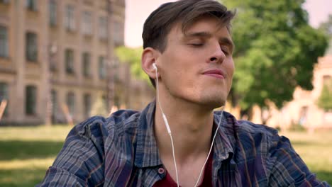 young handsome man is sitting on grass in park, listening music with earphones, singing, relax concept, building on background