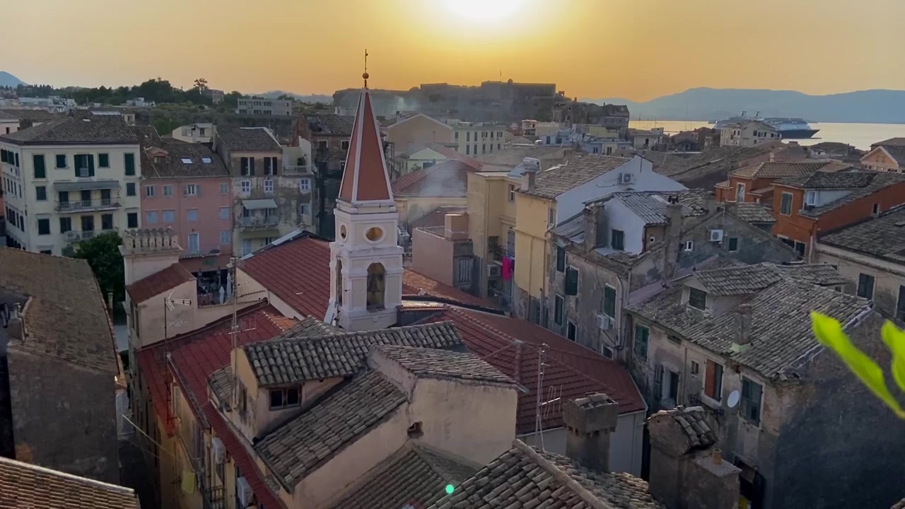 Vista del tramonto sulla Città Vecchia di Corfù, Grecia, che rivela un campanile in stile veneziano incorniciato dalla vegetazione in primo piano.