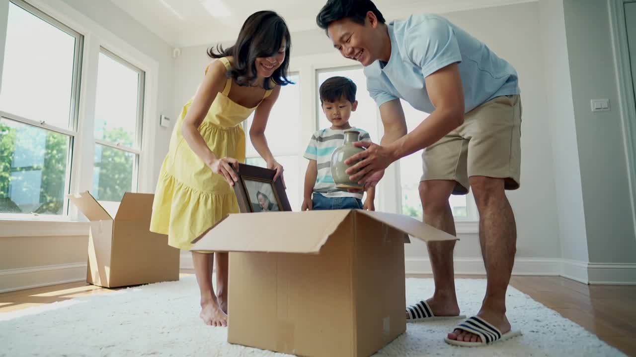 Opening box after move, mom in yellow dress lifting frame, dad handing vase, child pulling bear