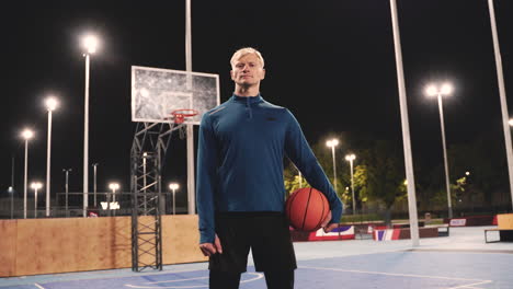 Jugador-De-Baloncesto-Confiado-Sosteniendo-La-Pelota-Y-Mirando-La-Cámara-Mientras-Está-De-Pie-En-Una-Cancha-Al-Aire-Libre-En-La-Noche-1