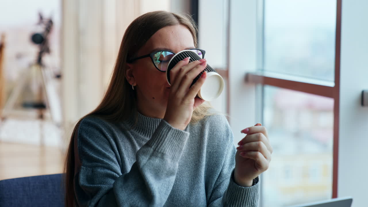 Smiling dreamy lady drinks coffee looking into window. Woman having a break from work at laptop. Lunch time at the working place. Close up.