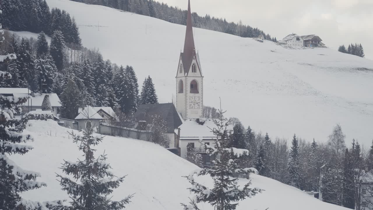 A red-steepled church stands in a snow-covered village at the foot of forested hills in South Tirol, Italy. Parallax video.
