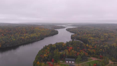 Pull-away-drone-shot-of-a-giant-twisting-river-surrounded-by-red,-orange,-and-yellow-leaf-trees-revealing-an-estate-on-the-waters-edge