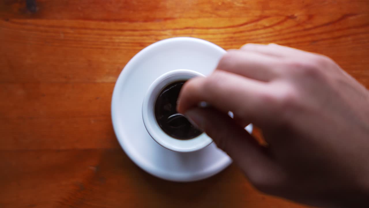 toma de una mano que está revolviendo azúcar en una taza de espresso caliente que está de pie sobre una mesa de madera marrón