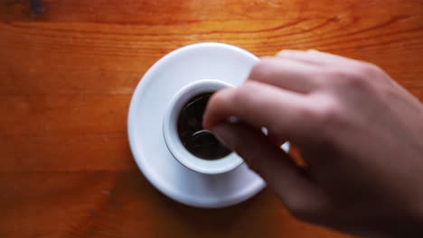 down shot of a hand that is stiring suger in a hot espress cup that is standing on a brown wooden table