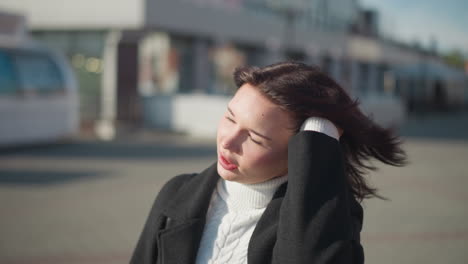 woman with brown hair in a black coat scratches her hair, displaying white polished nails on her left hand, against a sunny urban street backdrop with urban buildings