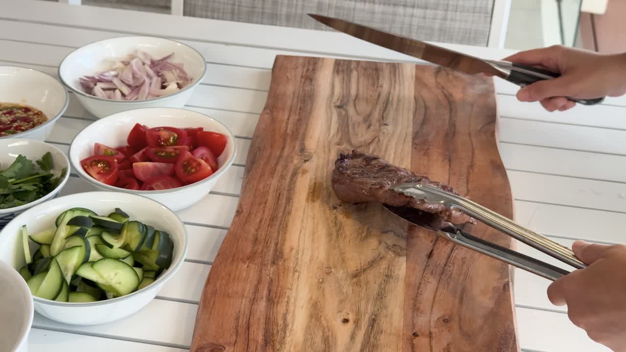 A person uses tongs and a knife to slice grilled beef steak on a wooden board, surrounded by bowls of fresh vegetables in bright natural light