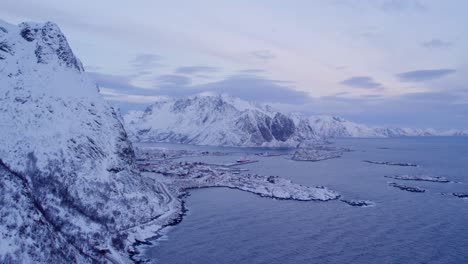 Impresionante-Vista-De-Lofoten-Con-Montañas-Cubiertas-De-Nieve,-Una-Serena-Masa-De-Agua-Y-Un-Pequeño-Asentamiento-Ubicado-Entre-Los-Elementos-Naturales.