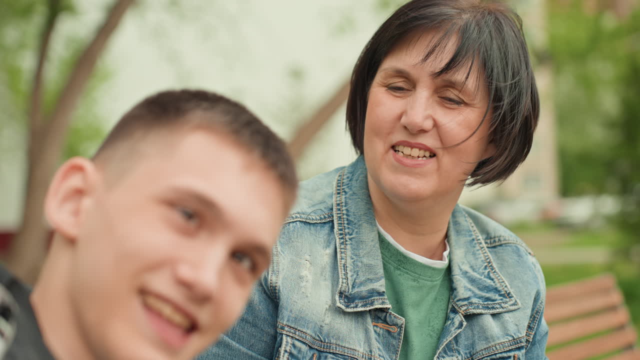 White Mother And Son Sitting On Bench In Park, Denim Jacket, Quiet Reflective Moment As Mother Watches Son With Gentle Concern Supportive Caregiver Bond, Thoughtful Expression, Calm Greenery
