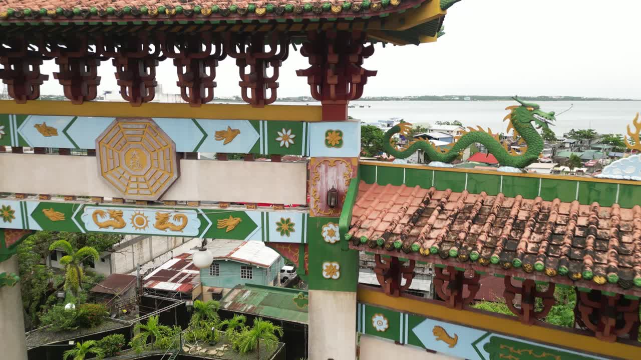 A high-angle wide shot shows Ma-Cho Temple’s ornate gate with curved eaves, dragon ornaments, and a golden emblem, framed by greenery and water in a calm, balanced scene