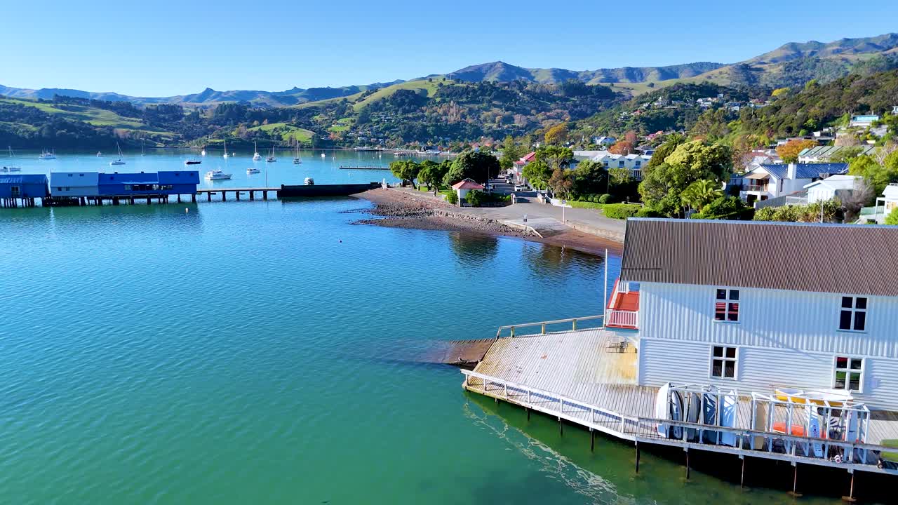 Aerial footage of Akaroa, showcasing vibrant waters, coastal buildings, and lush hills under clear skies