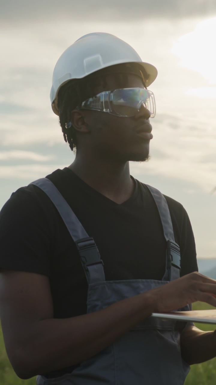 Man in hard hat and safety glasses uses tablet outdoors near wind turbines checking renewable energy
