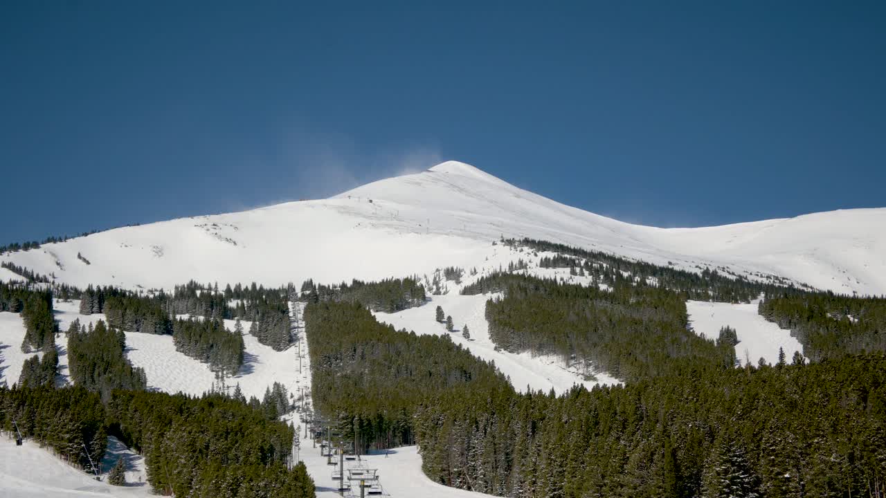 Snow blowing off the top of a mountain at a ski resort on a cloudless day, static