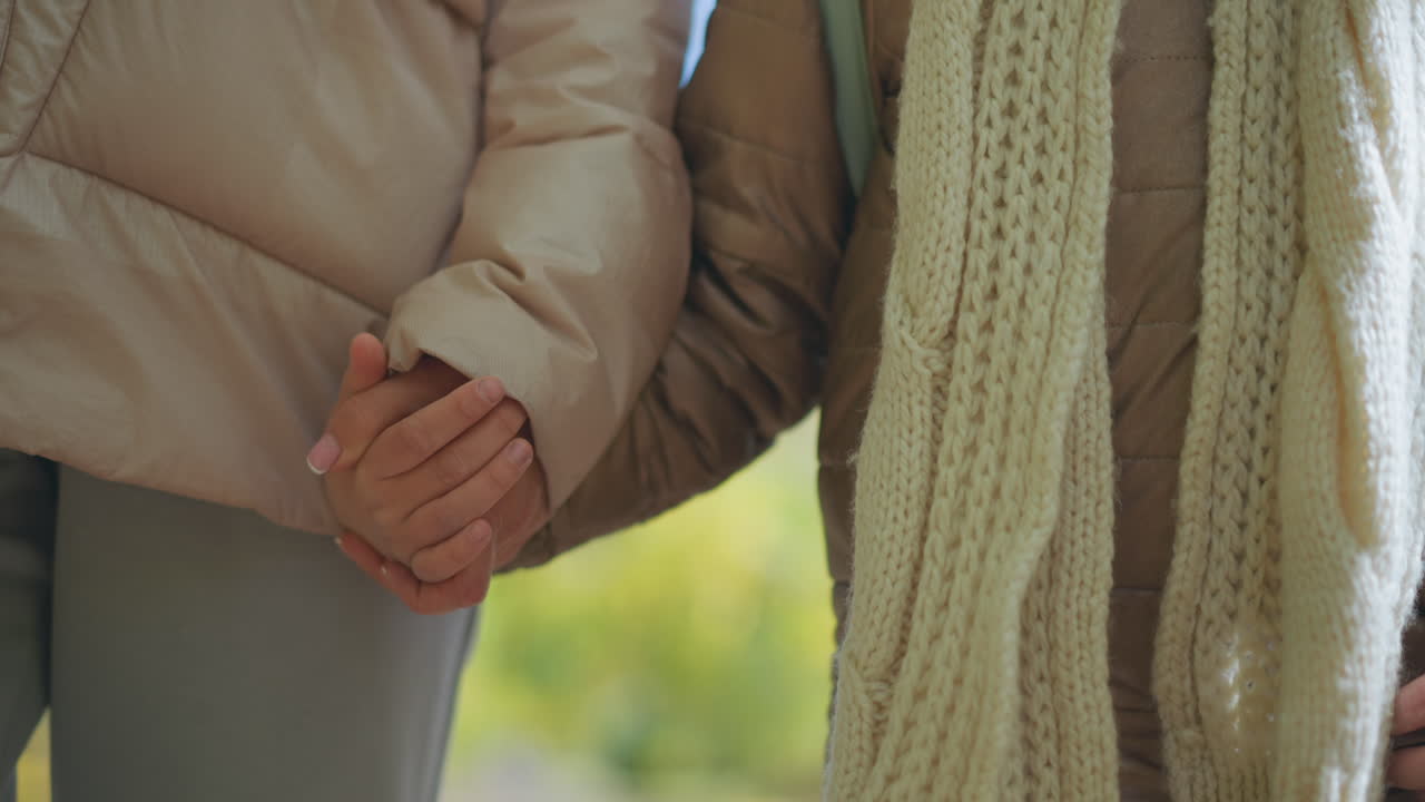 close up of adult and child interlocked hands walking through blurred park scene with green foliage and scattered dry leaves on ground wearing warm jackets and knit scarf