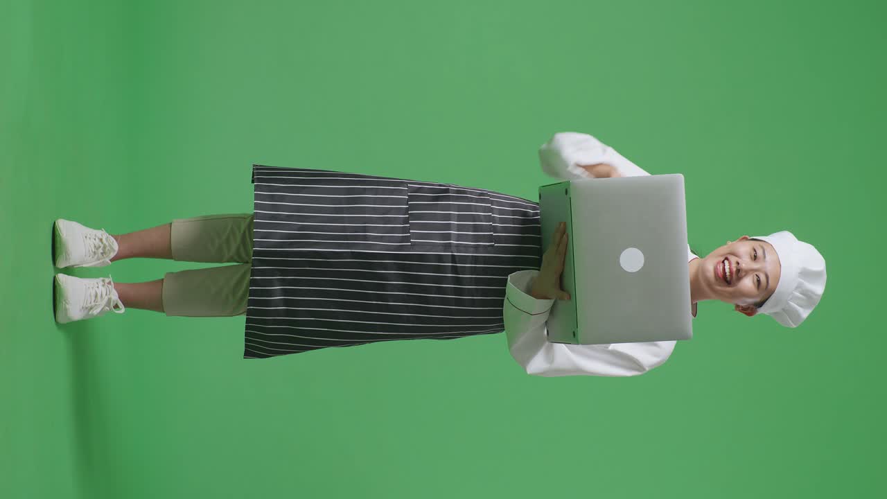 Full Body Of Asian Woman Chef Smiling And Pointing To A Laptop In Her Hand While Standing In The Green Screen Background Studio