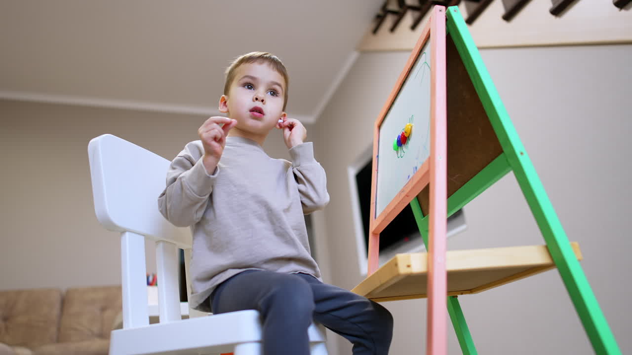 Lovely baby boy sits on the chair near the whiteboard. Low angle view at the child playing at home.