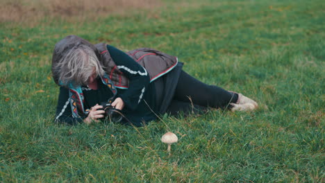 Ältere-Dicke-Frau-Mit-Grauen-Haaren,-Die-Auf-Dem-Gras-Liegt-Und-An-Einem-Kalten,-Windigen-Herbsttag-In-Zeitlupe-Einen-Sonnenschirmpilz-Fotografiert