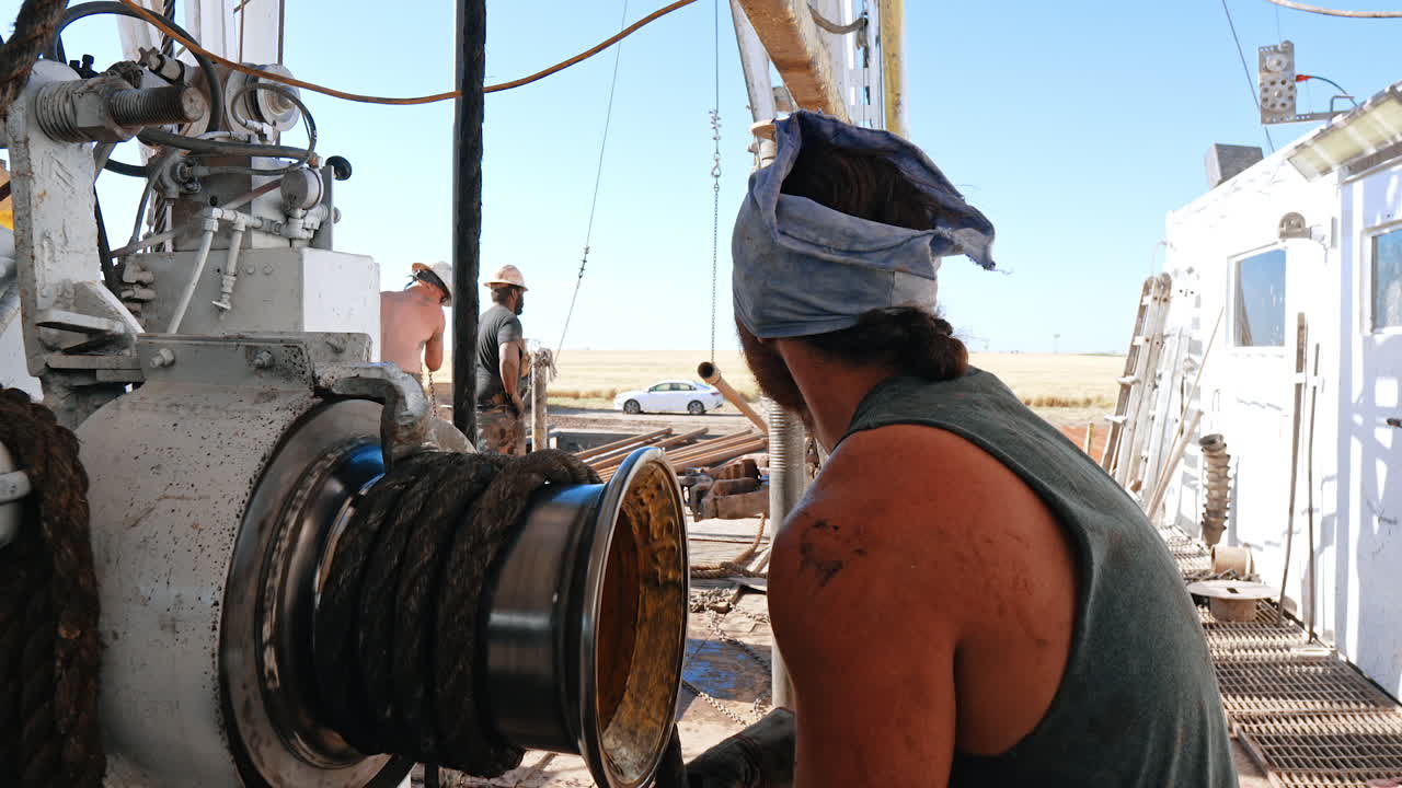 Rear view of a man winding the rope on the roller. Male workers setting equipment on the site for drilling oil or gas.