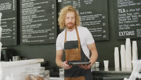 Porträt-Eines-Glücklichen-Kaukasischen-Männlichen-Baristas,-Der-Hinter-Der-Theke-Im-Café-Ein-Tablet-Benutzt