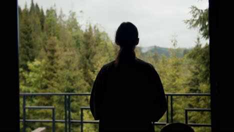 A-happy-brunette-girl-in-a-green-jacket-with-a-cup-of-tea-goes-to-the-balcony-of-a-country-house-overlooking-a-green-coniferous-forest-in-the-mountains