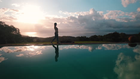 Woman-walks-in-slow-motion-next-to-a-calm-resort-pool,-her-silhouette-reflected-and-her-shadow-stretching-across-the-bottom-of-the-pool