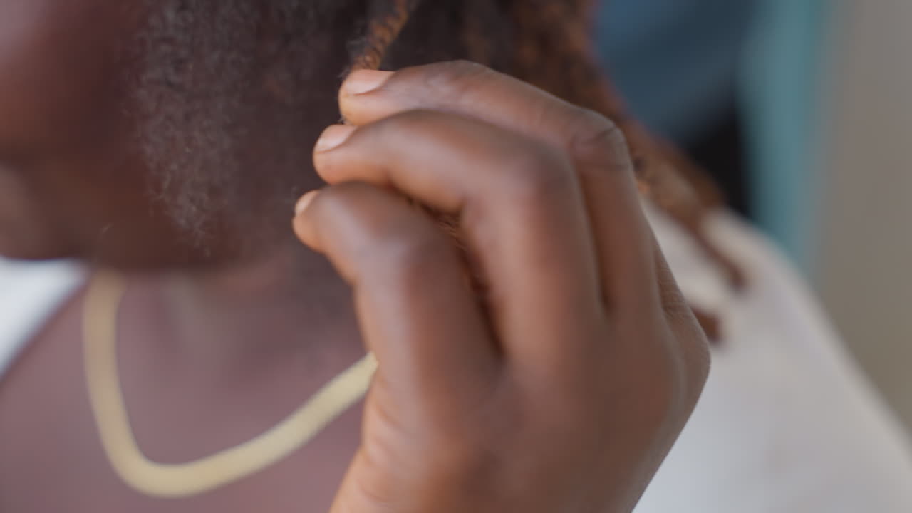 Black woman braiding hair with hands, closeup fingers twisting thin braids and gold necklace visible, natural texture and scalp detail in soft lighting, intimate home styling ritual, patient calm