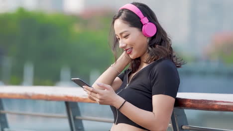 Portrait-active-girl-wearing-pink-headphones-sportwear-excercising-listening-to-music-leaning-on-railing-and-typing-smartphone