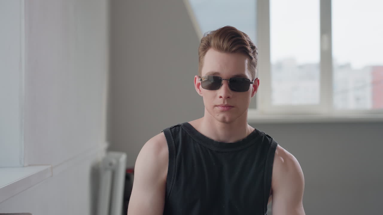 Young man with toned arms in black sleeveless shirt sits indoors near window, turning head to side with confident pose, bright daylight fills minimalist interior