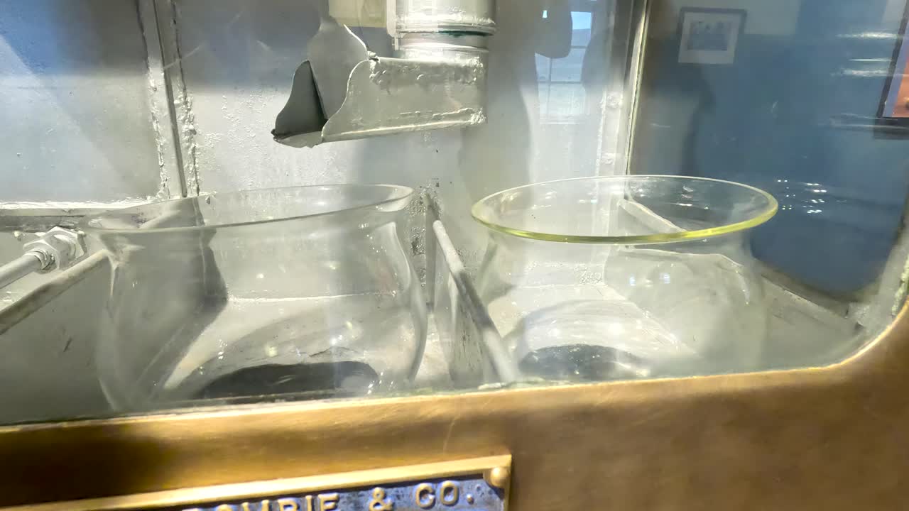 Clear liquid pours from metal spout into glass vessel inside distillery spirit safe, warm lighting