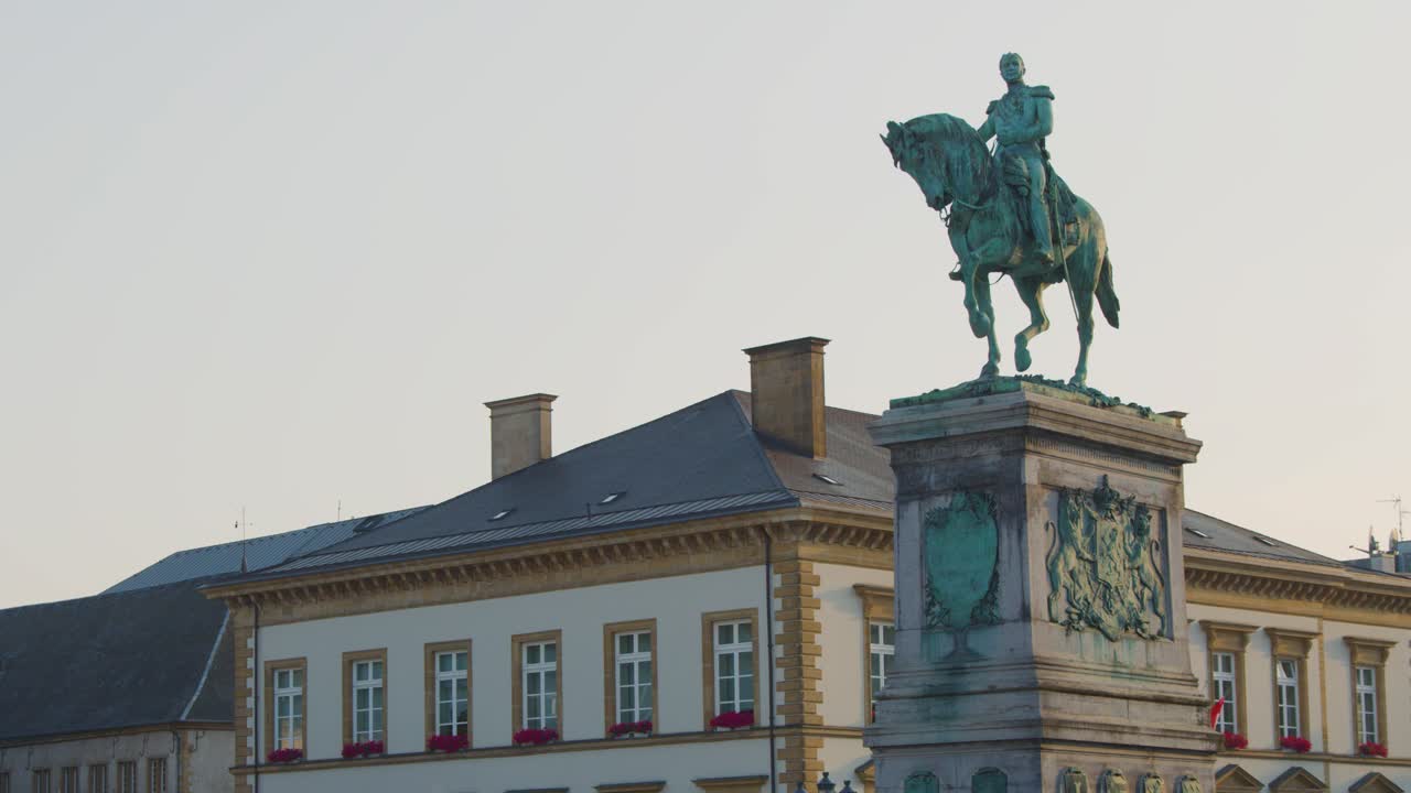 Camera slowly pans right, revealing bronze equestrian statue atop pedestal in urban square at dusk