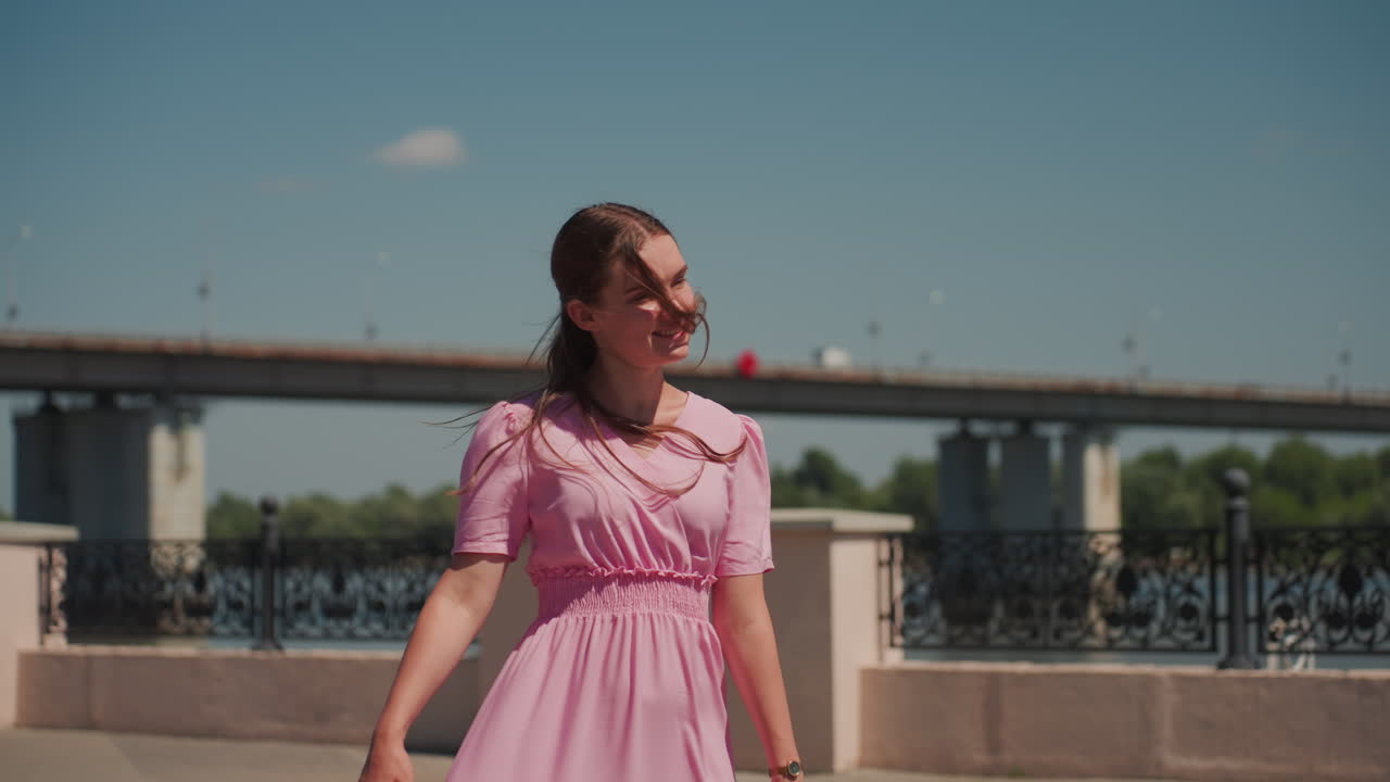Junge kaukasische Frau in rosa Kleid, die auf einer städtischen Uferpromenade spaziert, Rückansicht, weiches Sonnenlicht, geflochtene Haare, alte Laternenpfähle, Brücke im Hintergrund, dezentes Lächeln, cineastischer Sommer