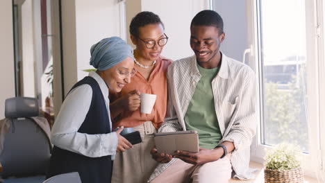 Eine-Muslimische-Geschäftsfrau,-Eine-Afroamerikanische-Geschäftsfrau-Und-Ein-Junger-Afrikanischer-Arbeiter-Lachen,-Während-Sie-Im-Büro-Etwas-Auf-Ein-Tablet-Schauen