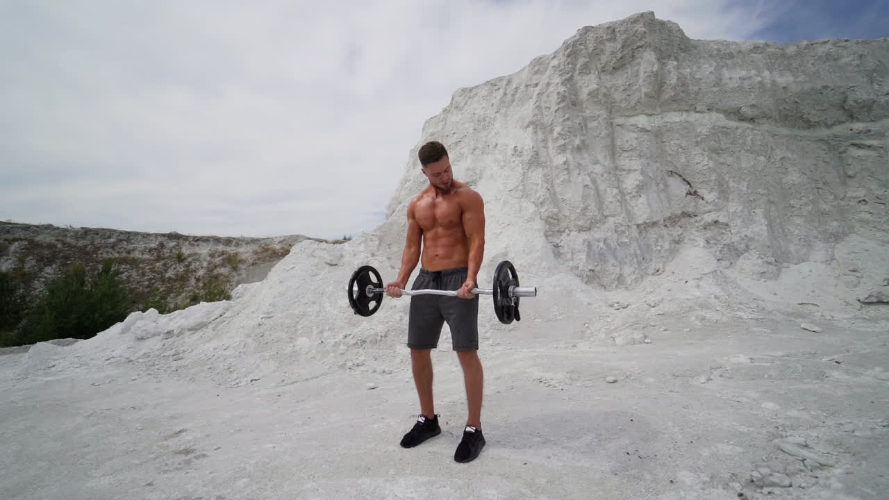 Handsome athlete training outdoors. Strong shirtless man with muscular body is lifting barbell on the white mountain background in summer. Slow motion.