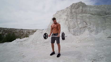 handsome athlete training outdoors. strong shirtless man with muscular body is lifting barbell on the white mountain background in summer. slow motion.