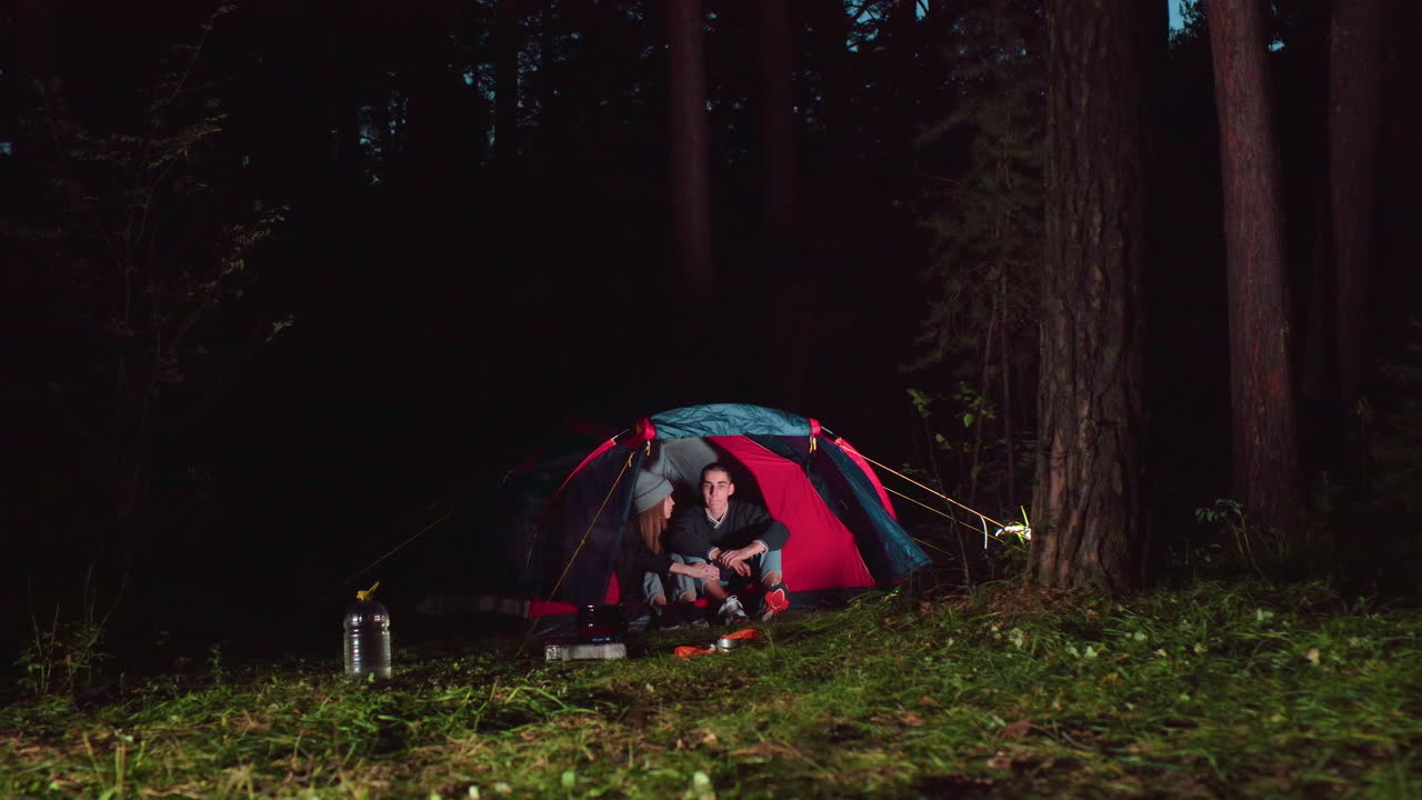 Lady gently rests head on boyfriend shoulder inside tent during calm evening in forest, capturing intimate and peaceful camping moment with warm lighting and serene natural surroundings