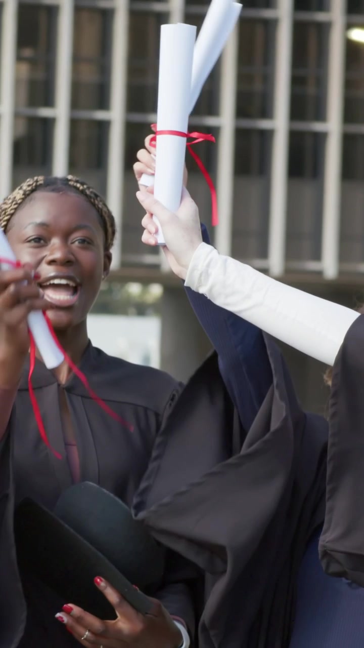 Vertical video: Camera widening, main grad in gown and friends raising diplomas on campus, cheering