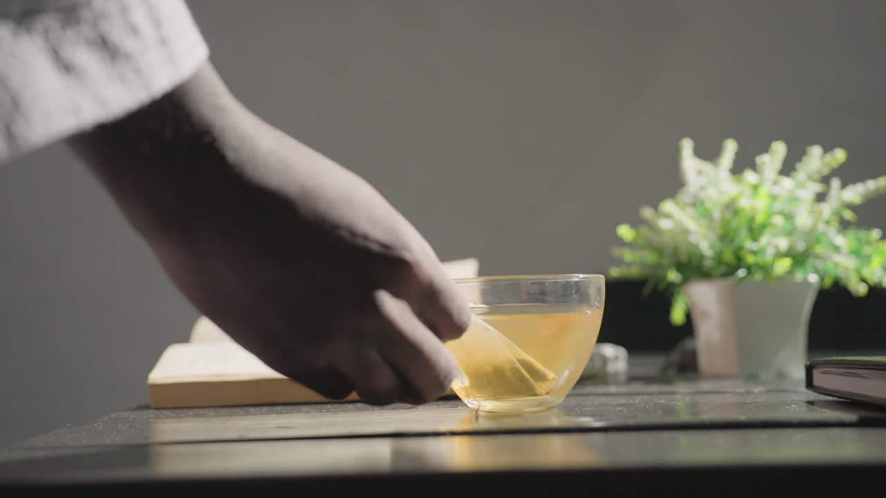 Person in bathrobe sitting at table, holding glass cup with tea, reading book, relaxing at home in modern cozy living room, enjoying leisure morning, healthy lifestyle concept