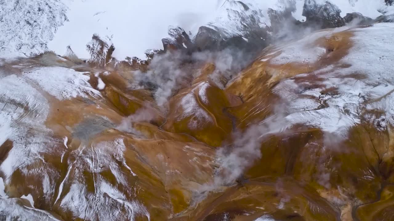Gorgeous aerial drone footage of brown, yellow and snow covered hot springs that are steaming in Iceland or Greenland during a clear day. Mountains are covered in white ice and snow, ground visible.