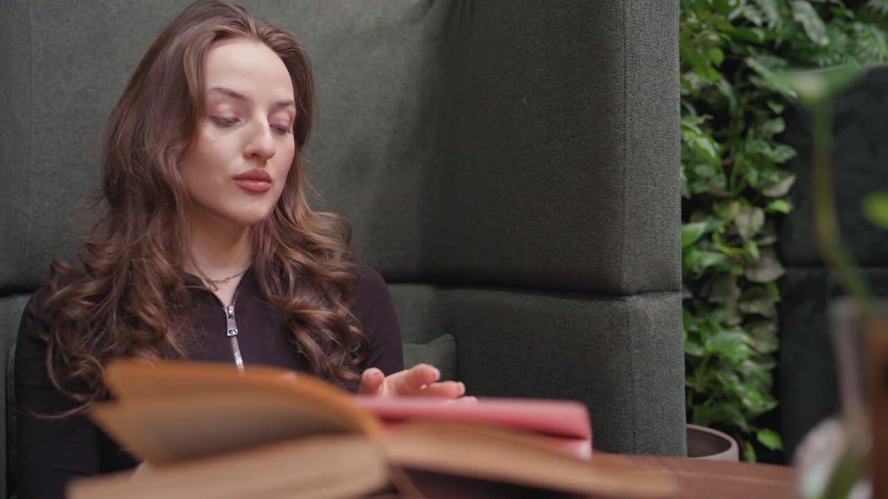 Lady seated in green booth pulls up sleeve with focused expression while preparing to write in open book on table, surrounded by cozy indoor environment and soft lighting