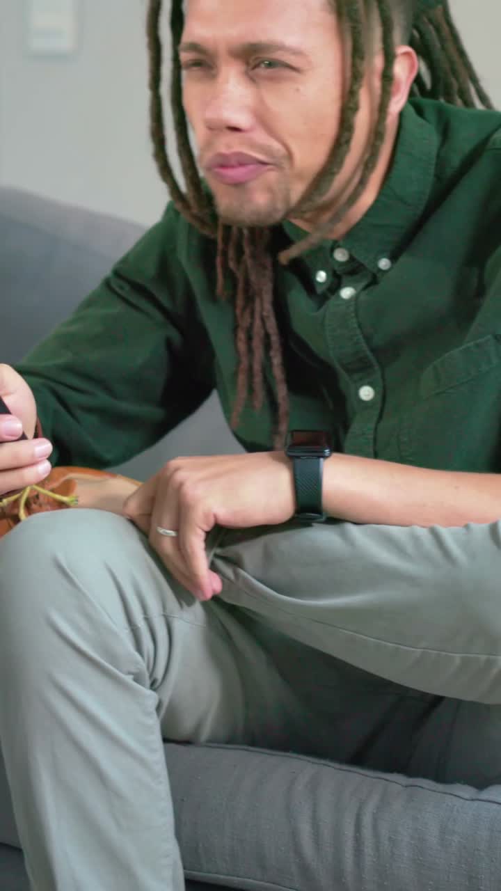 Video verticale: Uomo in maglietta verde che indica e preme il telecomando verso la TV, seduto su un divano grigio, mentre seleziona un programma.