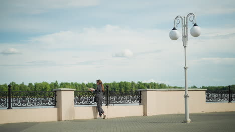 back view of a lady in grey clothing leaning against an iron railing while reading a book by the riverfront, with trees visible in the distance and a lamp post nearby