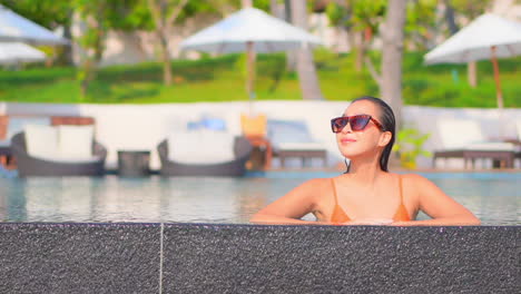 RIch-woman-relaxing-in-luxury-hotel-lounge-inside-swimming-pool-leaning-on-the-edge,-line-of-umbrellas-and-deckchairs-blurred-on-background,-slow-motion