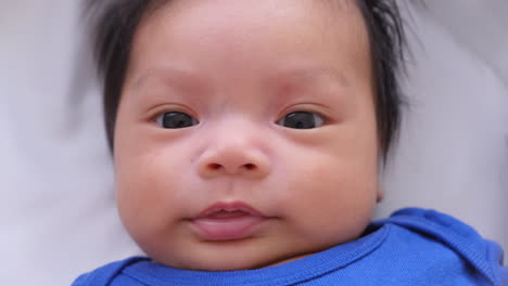 Angelic-Face-Of-Little-Baby-In-Blue-Onesie-Lying-In-Bed-And-Looking-At-Camera