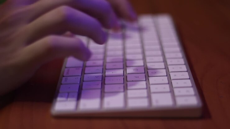 Caucasian hands in close up, typing on the white keys of a silver, wireless keyboard which is on a wooden table