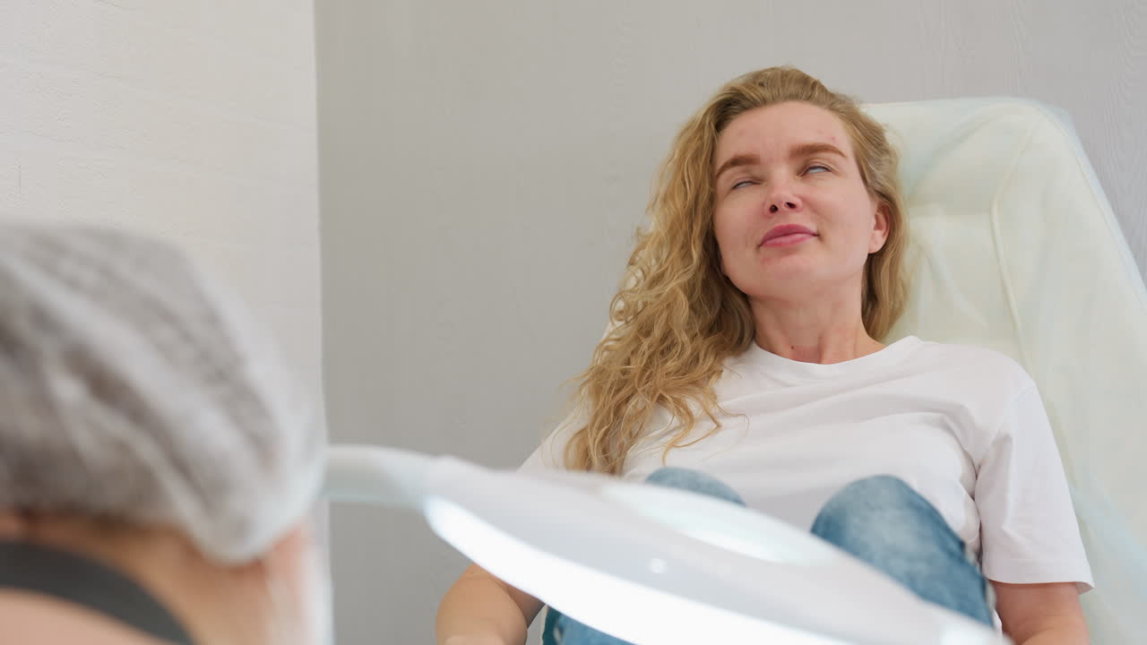 Client in white top sits on clinic chair gazing calmly at technician before gently closing eyes, expressing comfort, trust, and relaxation in clean, professional environment