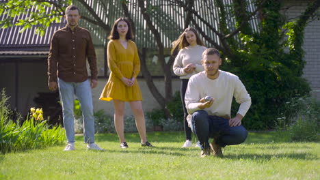 front view of caucasian young man squatting and throwing a petanque ball in the park on a sunny day while his friends waiting their turns