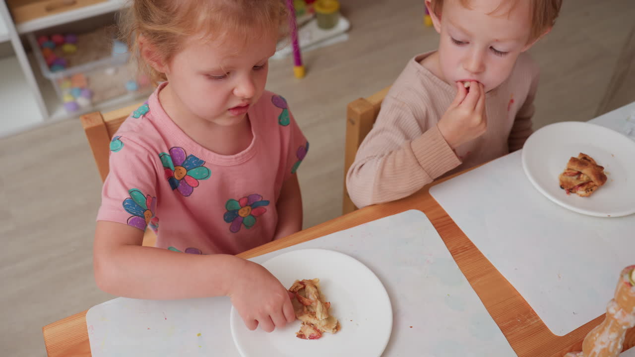 Bambina piccola esita con il dessert, solleva una fetta di torta di mele per ispezionarla mentre il bambino mangia la sua porzione e guarda, reazioni contrastanti durante la merenda all'asilo al tavolo, attenzione al gusto difficile e alla curiosità