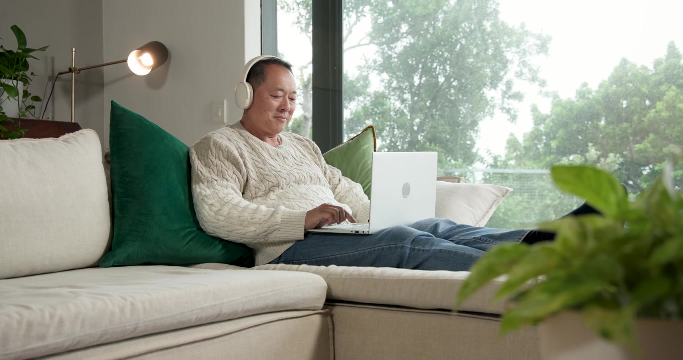 Anziano uomo asiatico che solleva l'auricolare, indossa le cuffie e digita su un laptop bianco lavorando da casa.