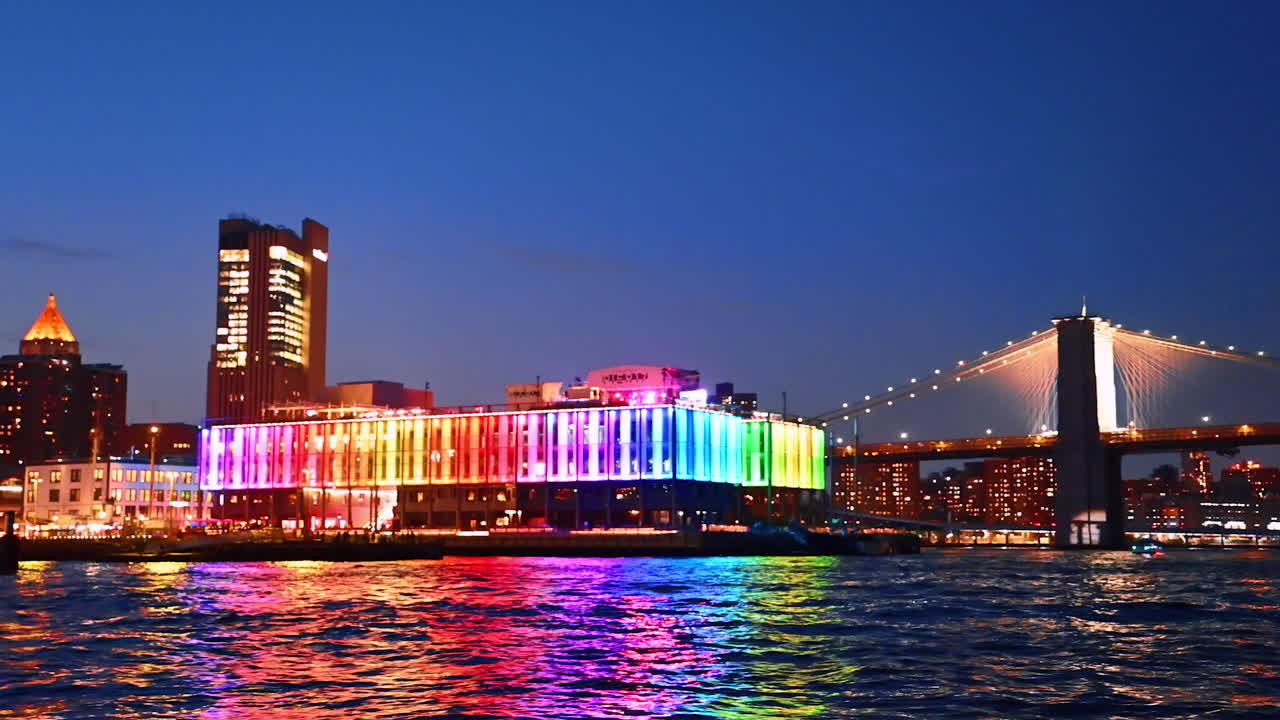 Rainbow illumination of Pier 17 Bar at night. The Brooklyn Bridge with street lights on at backdrop