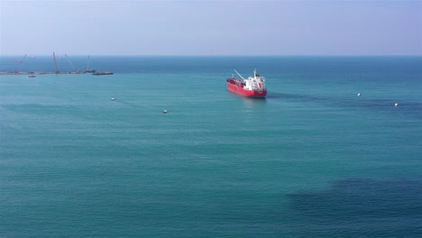 red cargo ship in calm blue sea with distant port activity